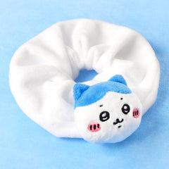 A white plush hair scrunchie featuring a cute cartoon cat face with blue ears, black eyes, and pink cheeks, set against a solid light blue background.